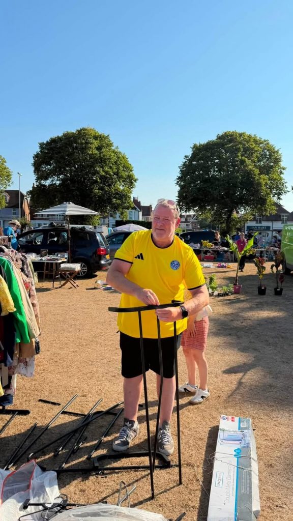 League Chairman Martin in a yellow league t-shirt putting a clothes rail together with the car boot sale in the background