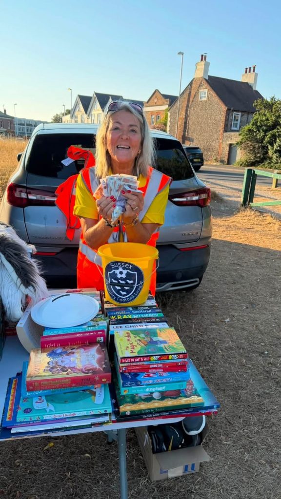 League Committee member Pauline in fron ot a car with a high vis jacket on a showing a handful of bank notes from her yellow collecting bucket
