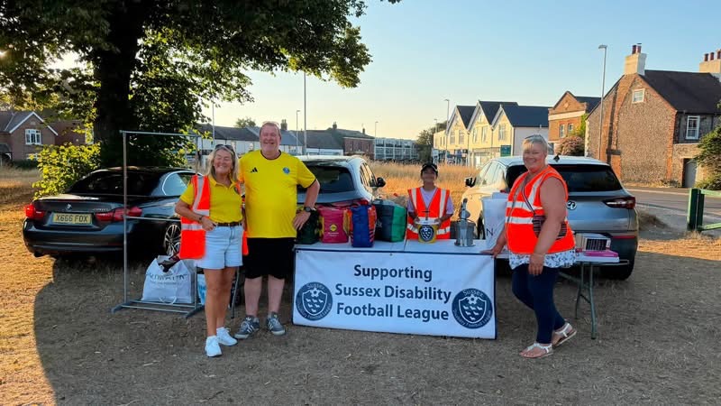 Four league helpers in high vis jackets standing by a table with the a banner in front saying Supporting Sussex Disability Football League. They are smiling and the sun is shining.
