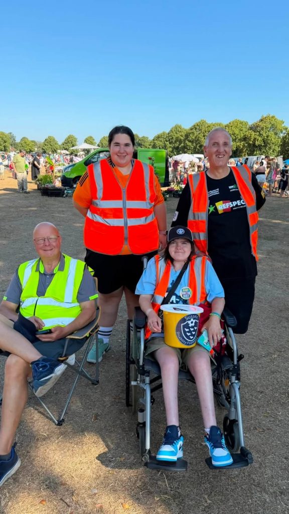 Four helpers in igh vis jackets. Two are standing, two are sitting, one in a chair and one in a wheelchair with a yellow collecting bucket with the league logo on the front